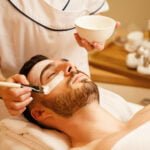 Unrecognizable beautician applying facial mask on man's face during spa treatment at wellness center.